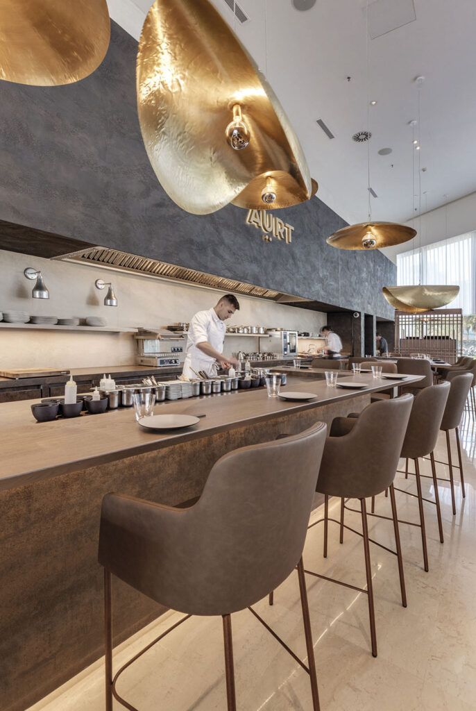 The interior of a contemporary restaurant showcasing a bar with a textured, dark brown Neolith Iron Corten countertop and matching front panel. Diners can sit on the brown bar stools and observe the chef preparing food in the brightly lit kitchen area behind the bar. Unique gold lighting fixtures add a warm glow to the scene.