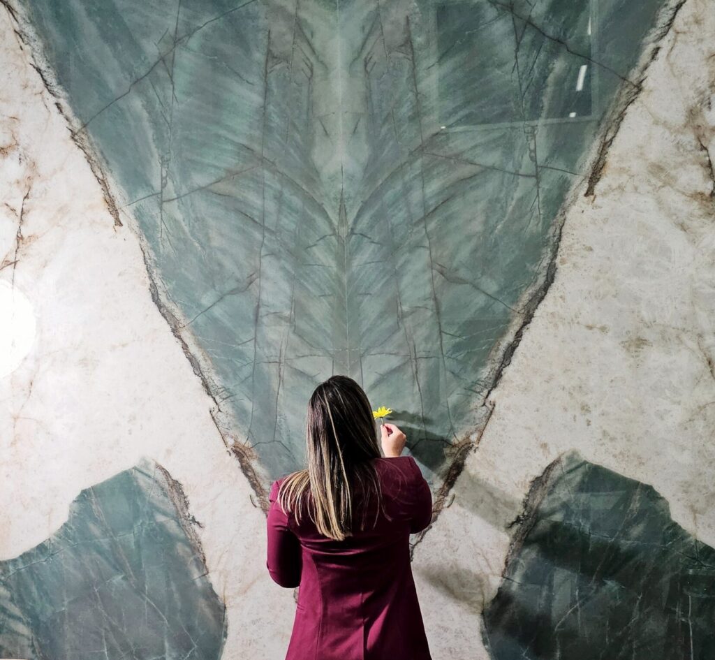 A person interacts with a large wall featuring a dramatic green and white veined pattern on a quartzite effect porcelain slab. They are holding a yellow flower.