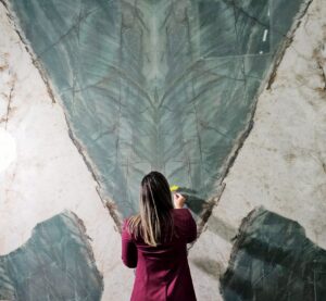 A person interacts with a large wall featuring a dramatic green and white veined pattern on a quartzite effect porcelain slab. They are holding a yellow flower.