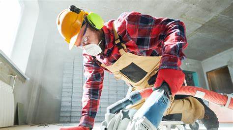 A construction worker wearing a dust mask and ear protection uses a power tool on a stone surface in an indoor construction site. Dust is visible in the air.